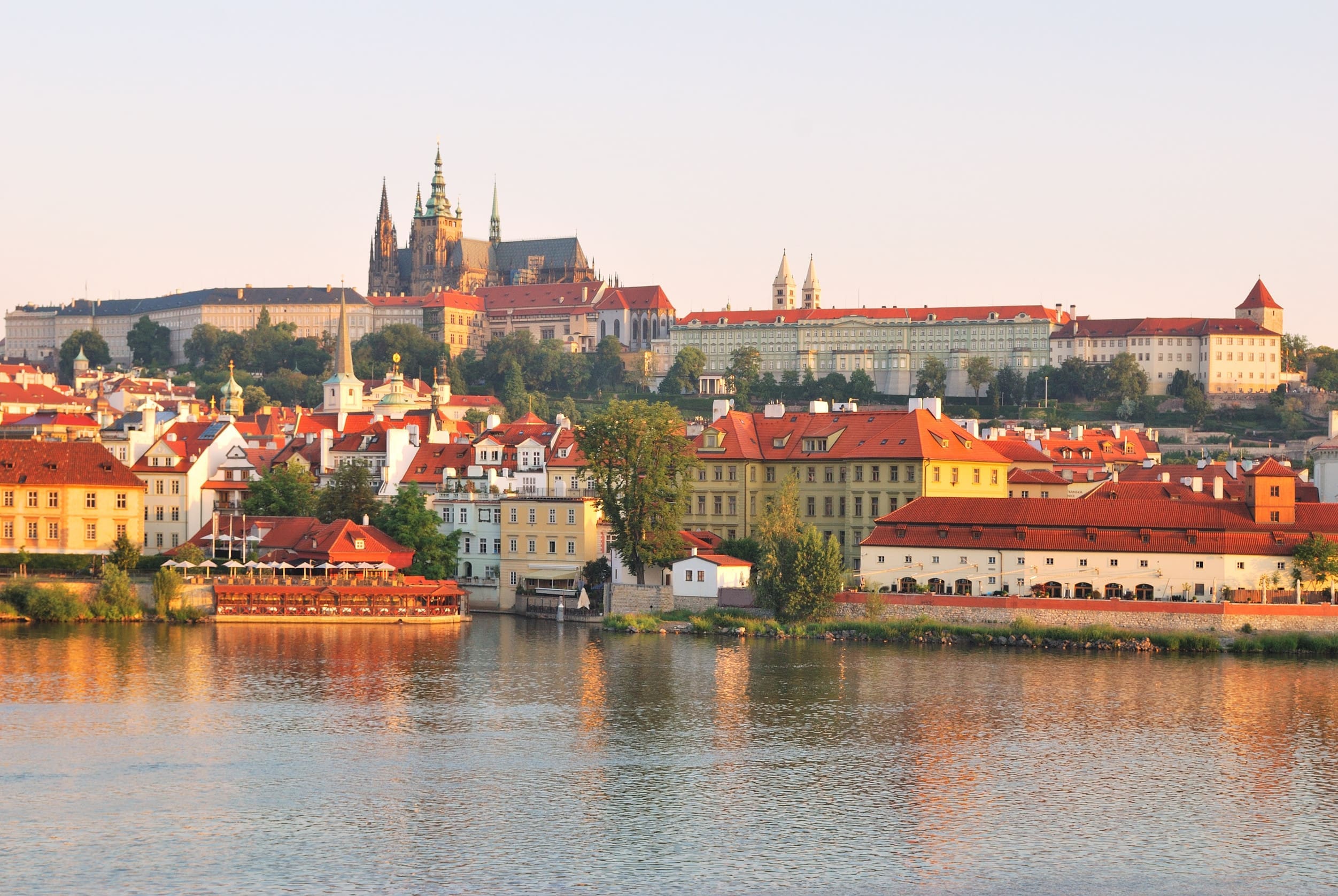 一張寬廣的風景照片，展示了捷克布拉格 (Prague) 沿著伏爾塔瓦河 (Vltava River) 的城市景觀和著名的布拉格城堡。