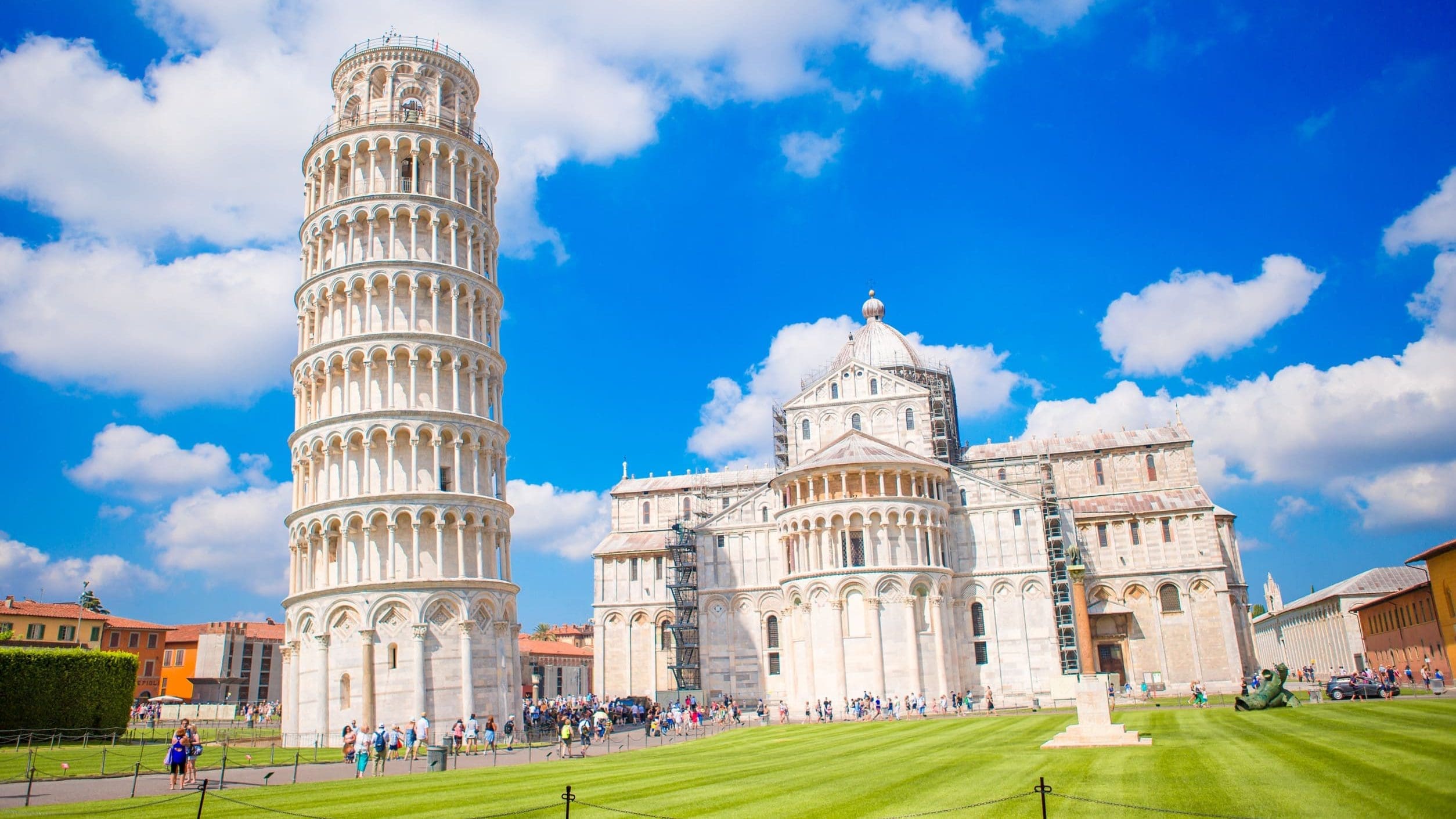 義大利比薩奇蹟廣場（Piazza dei Miracoli）的景觀。畫面左側是著名的比薩斜塔（Leaning Tower of Pisa），其傾斜的圓柱形身軀在藍天白雲下清晰可見。斜塔右側是比薩主教堂（Duomo di Pisa）的側面和圓頂。兩座建築均由白色大理石砌成，前方是修剪整齊的翠綠草坪，草坪上和周圍可見許多遊客。